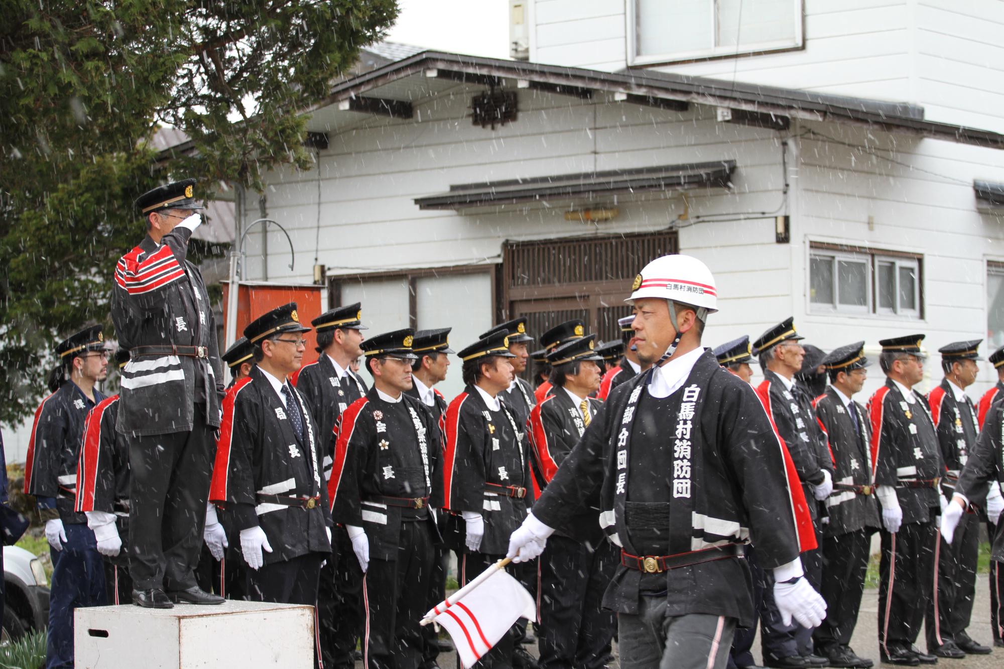 消防団に敬礼する村長と白い旗を持ちヘルメットをかぶった人が歩く分列行進の様子の写真